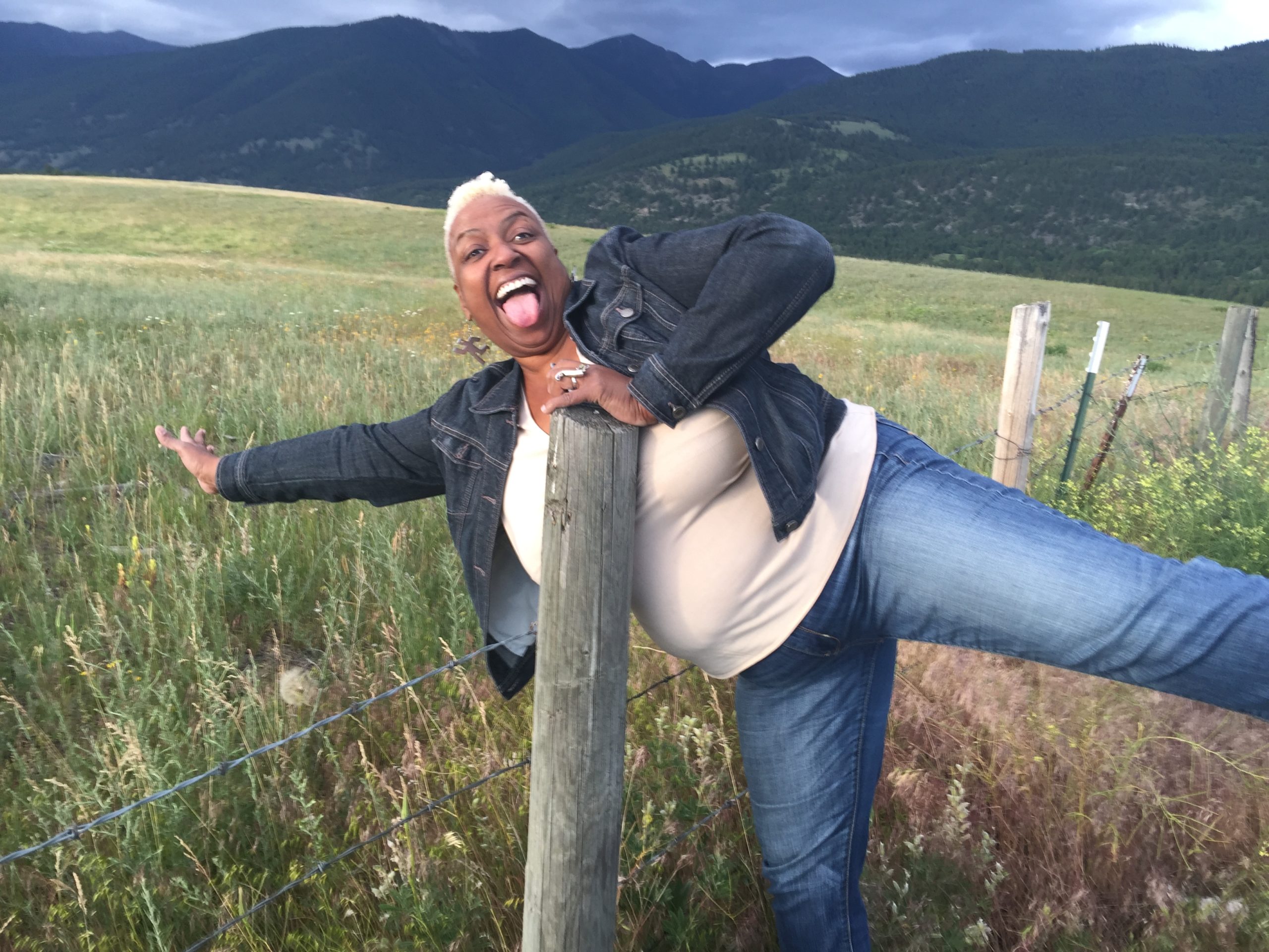 Christine Lincoln playful leans over a fence in a pasture, mountains in the background